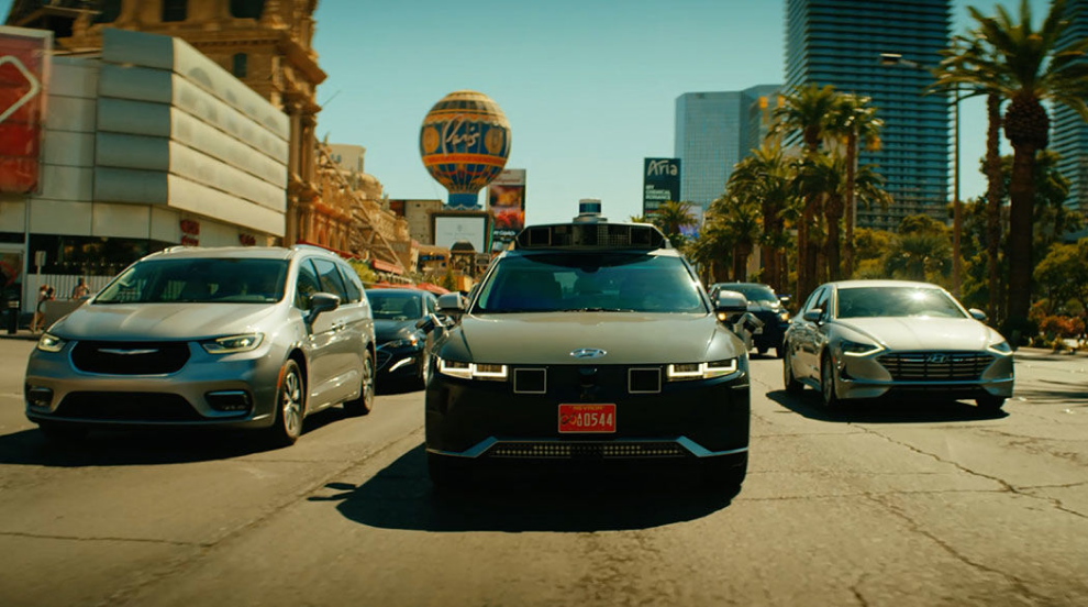灰色の IONIQ 5 ロボットタクシーが、晴れたラスベガスのダウンタウンで交通を運転しています。 太陽光がフロントガラスから反射していて、ロボットタクシーは中間車線にあります。